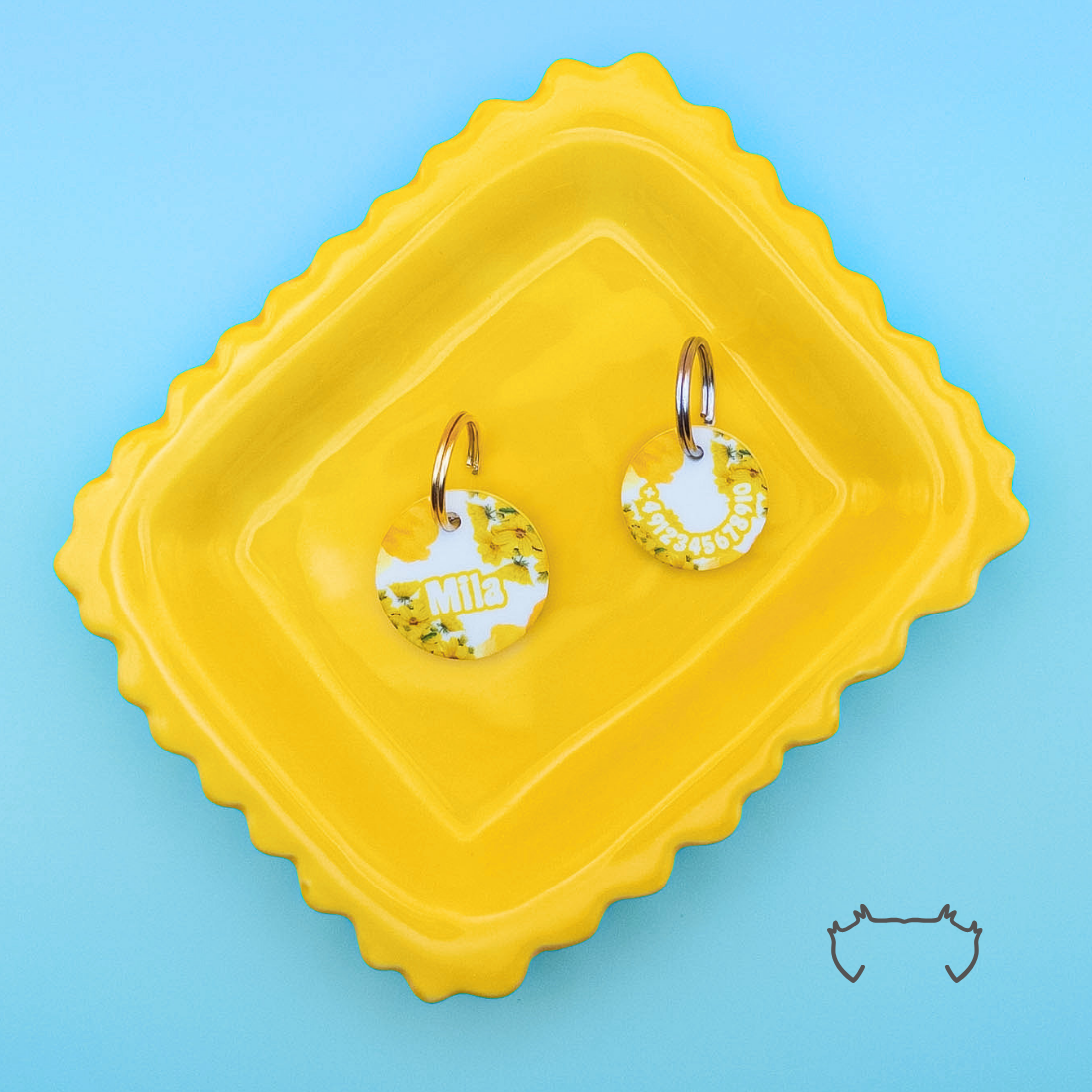 Zwei personalisierte runde Acryl-Hundemarken mit gelb-weißem Butterblumen-Design auf einem gelben Tablett vor türkisfarbenem Hintergrund.