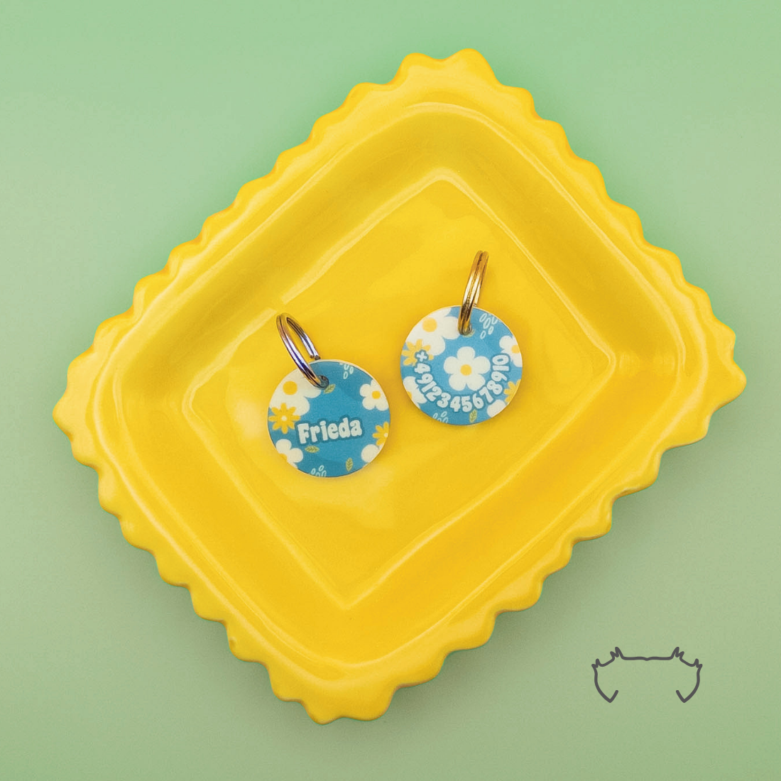 Zwei runde personalisierte Acryl-Hundemarken mit grün-gelbem Blumen-Design, Name und Telefonnummer, auf einem gelben Tablett vor hellgrünem Hintergrund.