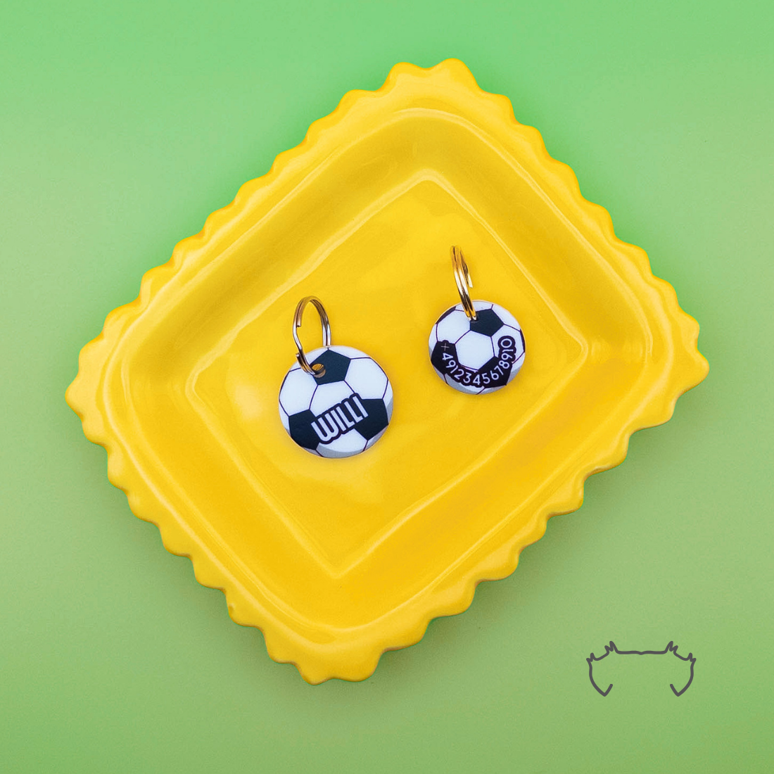 Zwei runde personalisierte Acryl-Hundemarken im Fußball-Design mit Name und Telefonnummer, auf einem gelben Tablett vor hellgrünem Hintergrund.