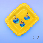Zwei runde Acryl-Hundemarken mit blau-grünem und orangefarbenem Gradient-Design, Name und Telefonnummer, auf einem gelben Tablett vor blauem Hintergrund.