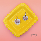 Zwei runde personalisierte Acryl-Hundemarken im hellen Luxury-Design mit zarten Beige- und Goldtönen auf einem gelben Tablett vor rosa Hintergrund, mit Name und Telefonnummer.