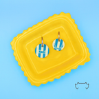 Zwei runde personalisierte Acryl-Hundemarken im petrol-beigen Streifen-Design auf einem gelben Tablett vor hellblauem Hintergrund, eine Hundemarke mit Name „Nami“ und eine mit Telefonnummer.