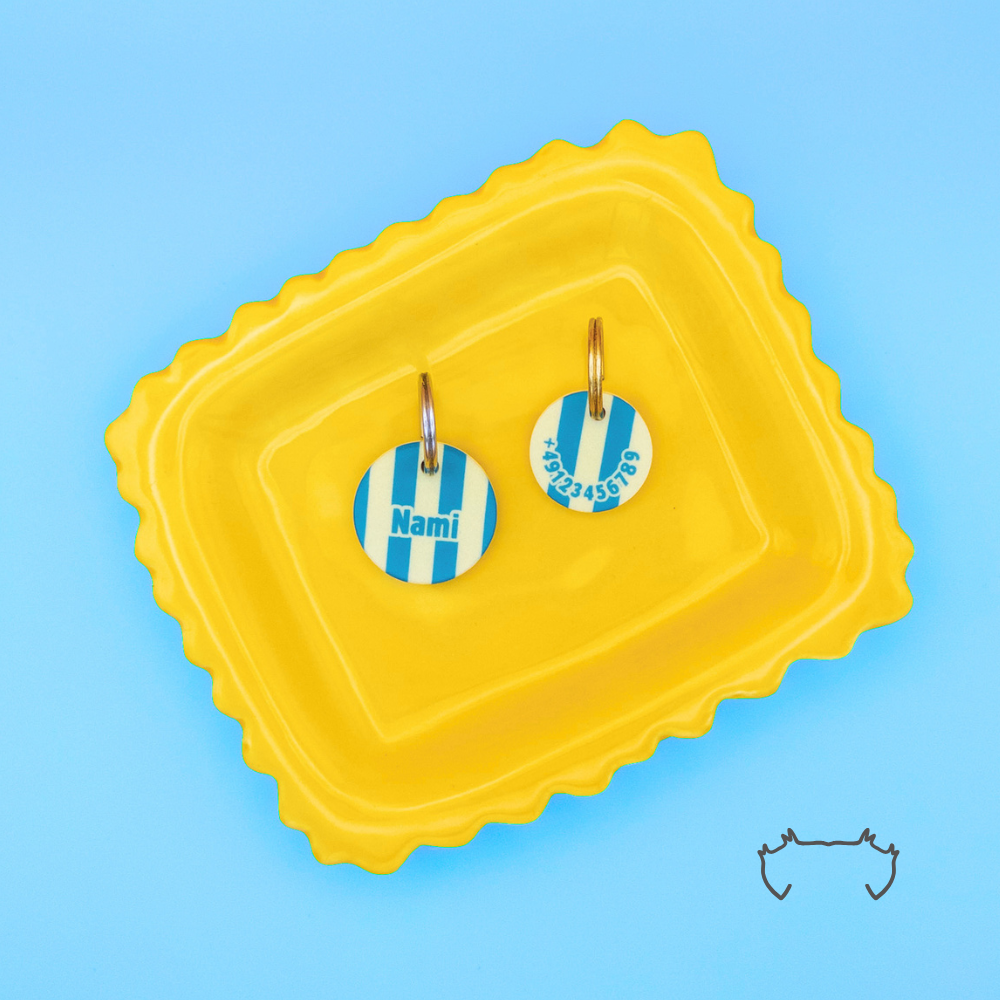Zwei runde personalisierte Acryl-Hundemarken im petrol-beigen Streifen-Design auf einem gelben Tablett vor hellblauem Hintergrund, eine Hundemarke mit Name „Nami“ und eine mit Telefonnummer.