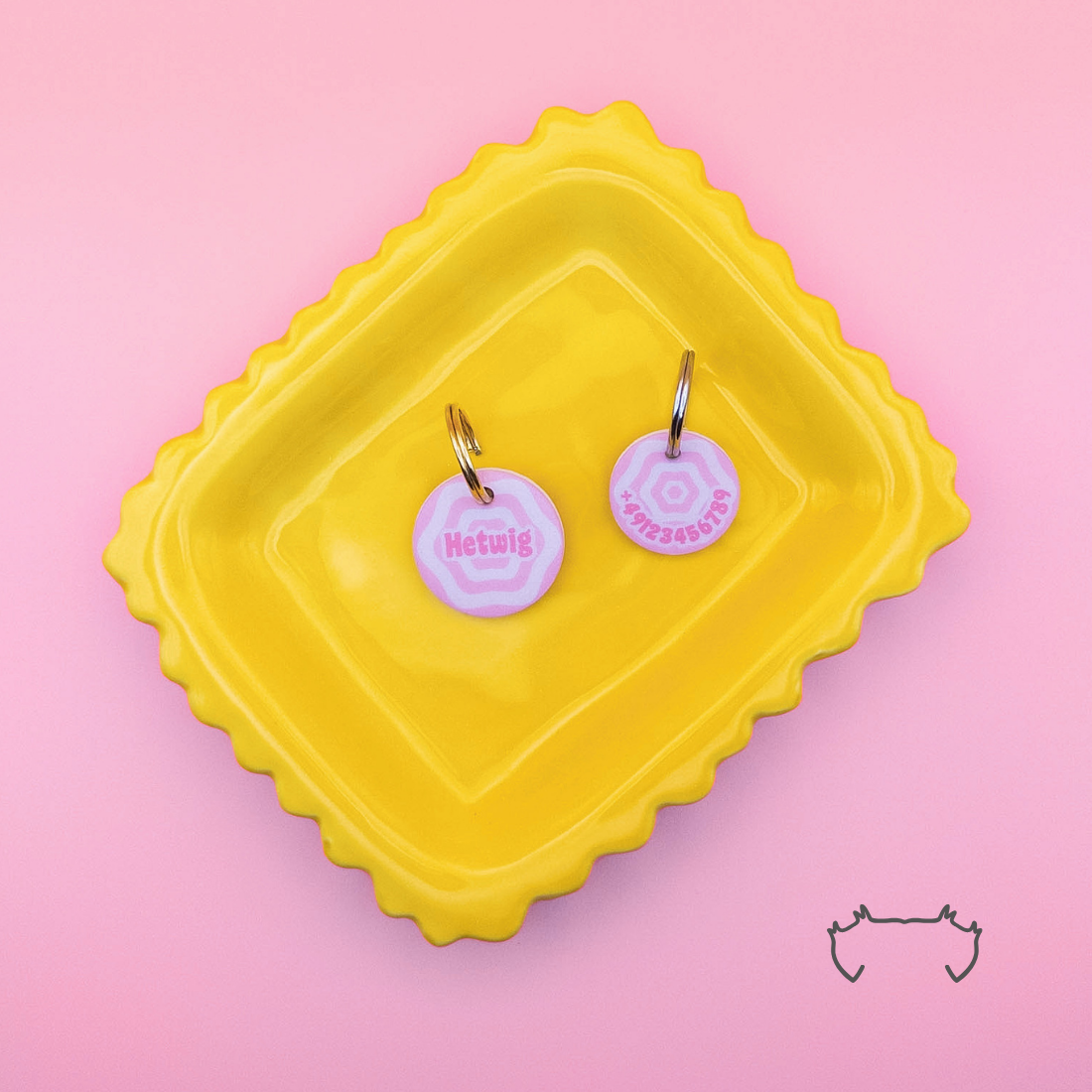 Zwei runde Acryl-Hundemarken im rosa Y2K-Design mit spiralförmigem Muster, Name und Telefonnummer, auf einem gelben Tablett vor rosa Hintergrund.