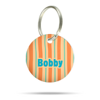 Personalisierte runde Acryl-Hundemarke für Hunde mit orangefarbenem, cremefarbenem und türkisfarbenem Streifenmuster, türkisblauem Namen „Bobby“ und silbernem Schlüsselring auf weißem Hintergrund.
