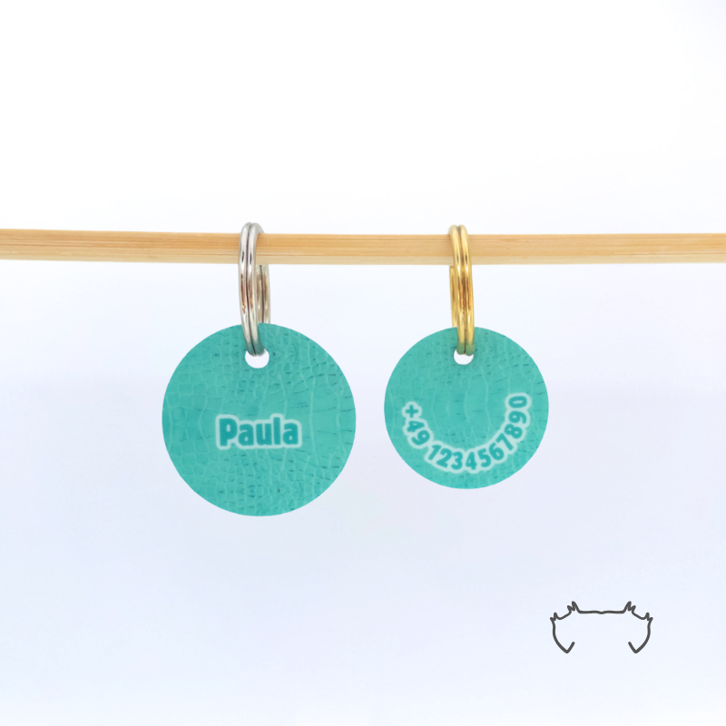 Zwei runde Acryl-Hundemarken im türkisfarbenen Croco-Design an einer hellen Holzstange, vorne mit Name „Paula“ und hinten mit Telefonnummer.