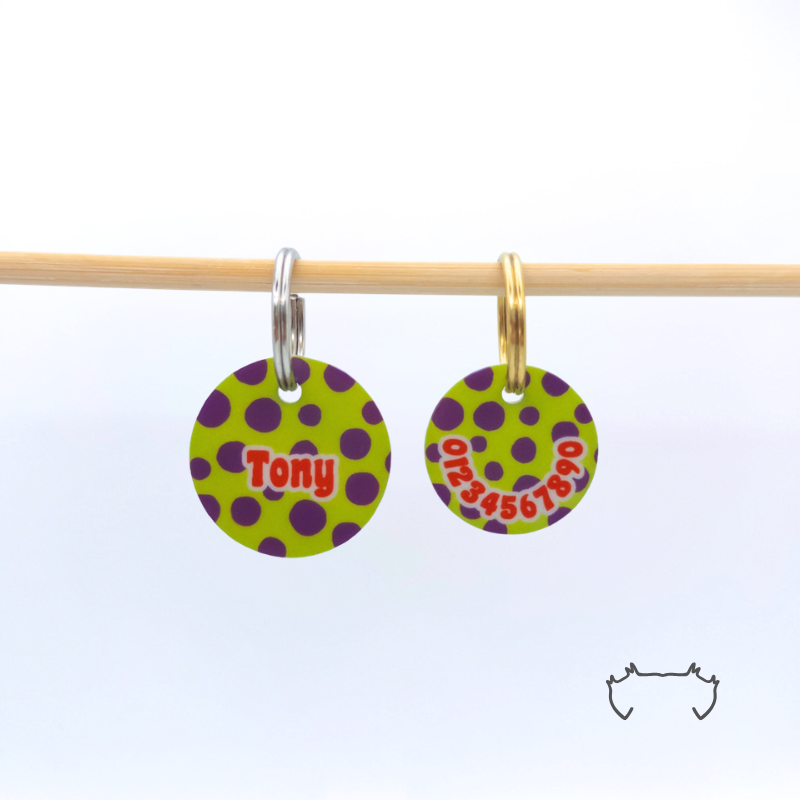 Zwei personalisierte runde Acryl-Hundemarken im grün-lila Punkte-Design an einer hellen Holzstange, vorne mit Name „Tony“ und hinten mit Telefonnummer.
