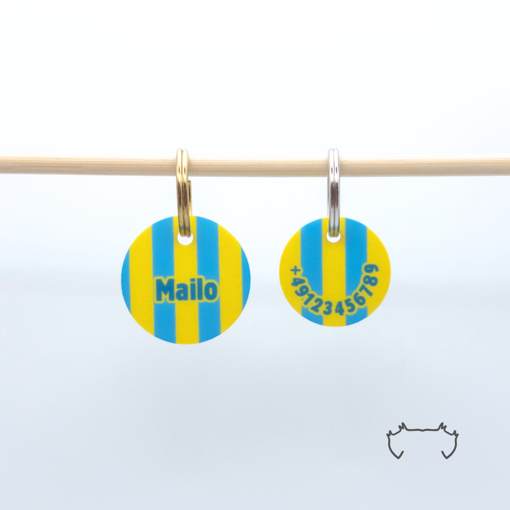 Zwei personalisierte runde Acryl-Hundemarken für Hunde im gelb-blauen Streifenmuster an einer hellen Holzstange, vorne mit Name „Mailo“ und hinten mit Telefonnummer.