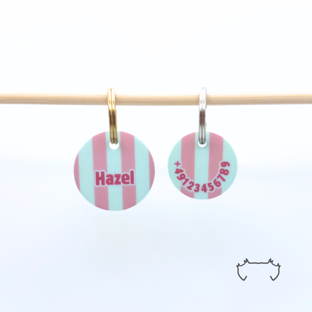 Zwei personalisierte runde Acryl-Hundemarken für Hunde im rosa, mintgrünen und weißen Streifenmuster an einer hellen Holzstange, links die Vorderseite mit Name „Hazel“, rechts die Rückseite mit Telefonnummer.