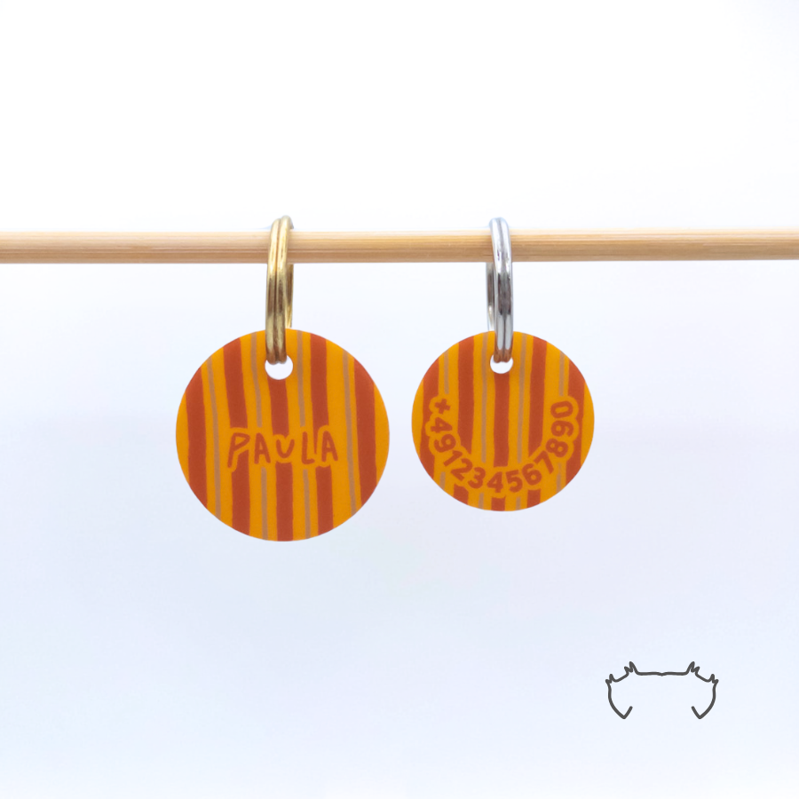Zwei runde personalisierte Acryl-Hundemarken mit orange-braunem Herbst-Streifenmuster an einer hellen Holzstange, eine Marke mit Name und eine mit Telefonnummer.