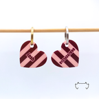 Zwei formgeschnittene Acryl-Hundemarken in Herzform an einer hellen Holzstange, mit bordeauxrotem, rosa und schwarzem Streifenmuster, eine Marke mit Name und eine mit Telefonnummer.