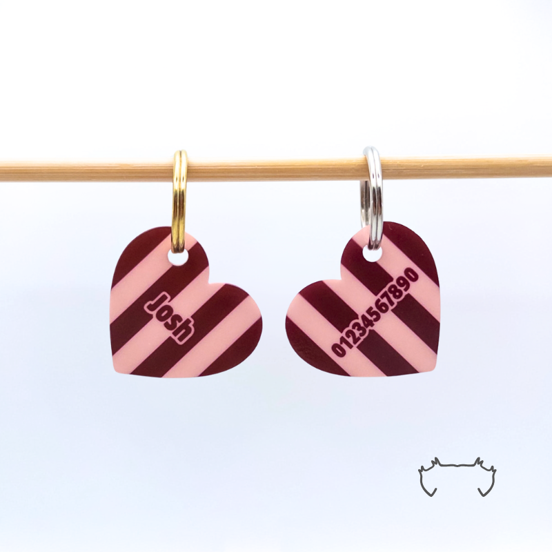 Zwei formgeschnittene Acryl-Hundemarken in Herzform an einer hellen Holzstange, mit bordeauxrotem, rosa und schwarzem Streifenmuster, eine Marke mit Name und eine mit Telefonnummer.