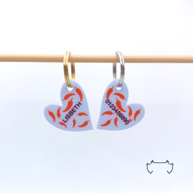 Zwei leichte, formgeschnittene Acryl-Hundemarken in Herzform an einer hellen Holzstange, mit hellblauem Design, roten Chili-Motiven, Name „LISBETH“ und Telefonnummer auf der Rückseite.