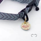 Runde Acryl-Hundemarke mit beigem Blumenmuster und personalisiertem Namen, befestigt an einem grauen Hundehalsband mit dunklem Chevron-Muster und schwarzem Verschluss.
