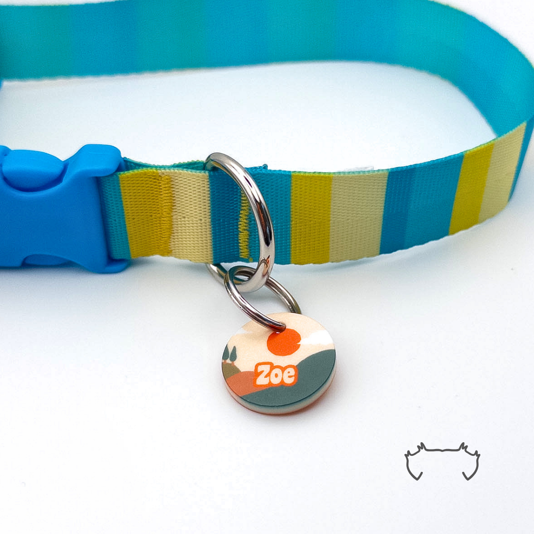 Personalisierte runde Acryl-Hundemarke mit Berg- und Sonnenaufgangsmotiv in Rot, Grün und Beige, befestigt an einem türkis-gelb-beige gestreiften Hundehalsband.