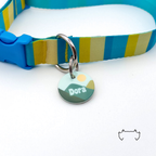 Personalisierte runde Acryl-Hundemarke mit grünem Bergmotiv und heller Sonne, befestigt an einem türkis-gelb-beige gestreiften Hundehalsband mit blauem Verschluss.