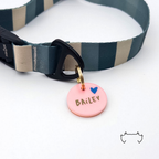 Runde personalisierte Acryl-Hundemarke in zartem Rosa mit Herzmotiv und Name, befestigt an einem dunkelgrün-beige gestreiften Hundehalsband mit schwarzem Verschluss.
