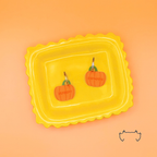Zwei bunte, formgeschnittene Acryl-Hundemarken in Kürbisform auf einem gelben Tablett vor orangefarbenem Hintergrund. Die leichten Hundemarken zeigen ein herbstliches Kürbis-Design mit Personalisierung.