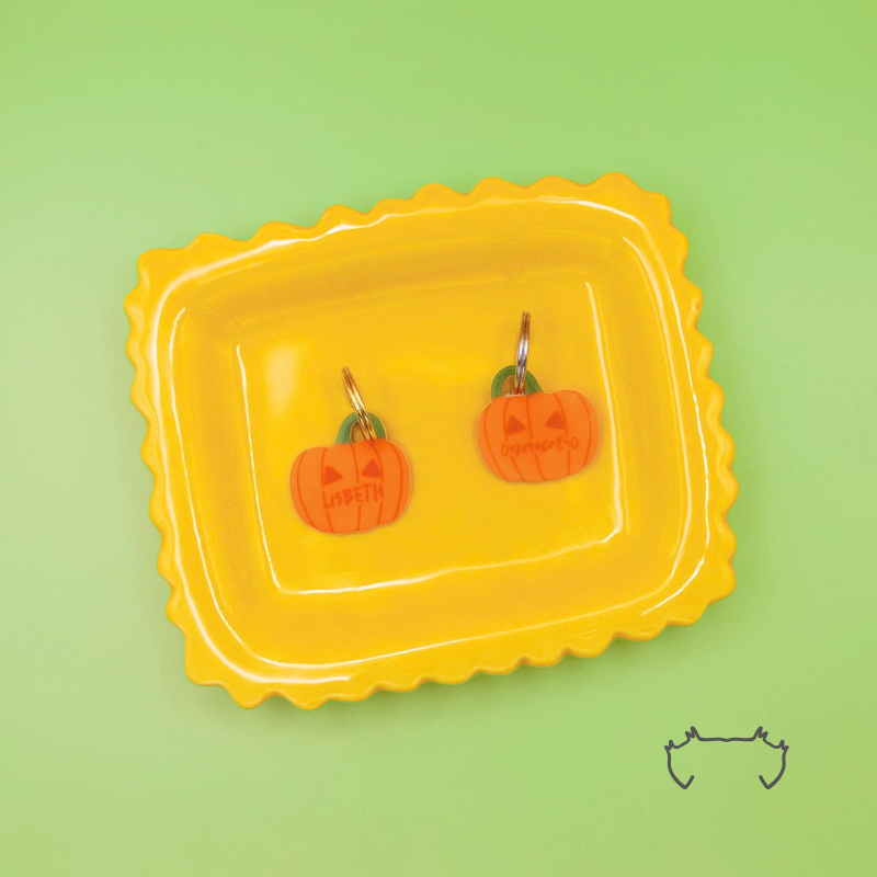 Zwei leichte, formgeschnittene Acryl-Hundemarken in Kürbisform auf einem gelben Tablett vor hellgrünem Hintergrund. Die bunten Hundemarken zeigen ein orangefarbenes Halloween-Kürbis-Design mit Personalisierung.