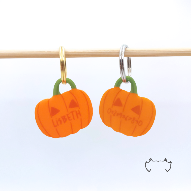 Zwei formgeschnittene Acryl-Hundemarken in Kürbisform an einer hellen Holzstange, mit orangefarbenem Halloween-Design, Name und Telefonnummer. Bunte leichte Hundemarke für Hunde mit herbstlichem Kürbis-Motiv.