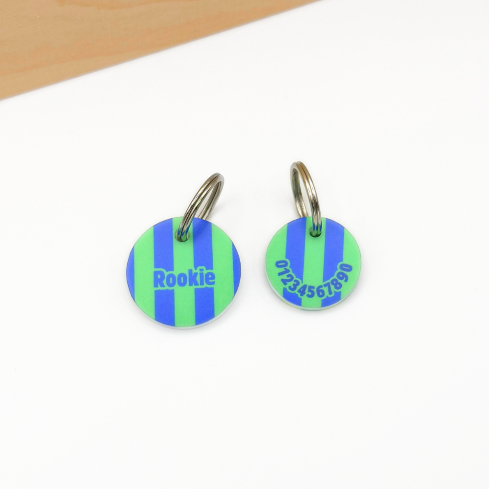 Zwei personalisierte Hundemarken aus Acryl mit blau-grünem Streifenmuster liegen auf einem weißen Untergrund. Links ist die Vorderseite mit dem Namen „Rookie“ zu sehen, rechts die Rückseite mit Telefonnummer. Beide Marken sind mit silberfarbenen Schlüsselringen versehen. Im oberen Bildbereich verläuft eine helle Holzleiste.