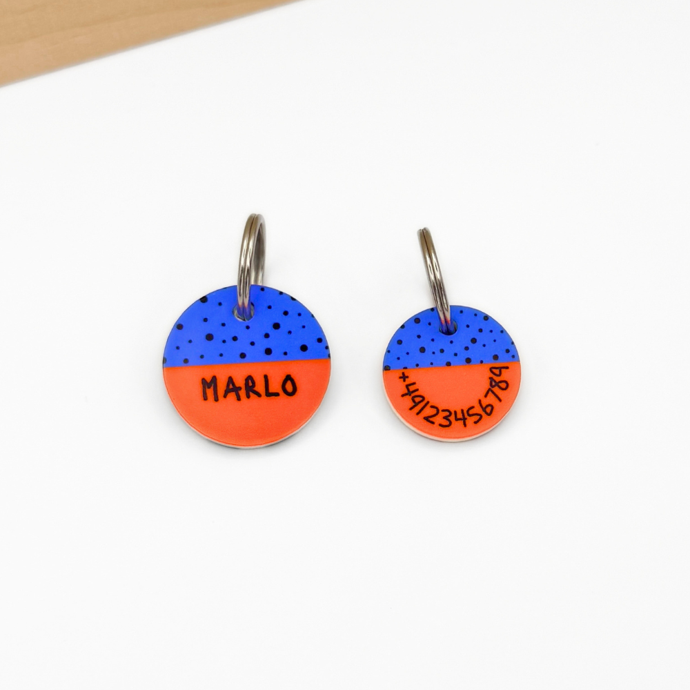 Zwei personalisierte Hundemarken aus Acryl auf weißem Untergrund. Die linke Marke zeigt den Namen „Marlo“ auf einem blau-orangen Design mit schwarzem Punktmuster. Rechts liegt die Rückseite mit aufgedruckter Telefonnummer. Beide Marken sind an silberfarbenen Metallringen befestigt. Im Hintergrund ist eine helle Holzleiste sichtbar.