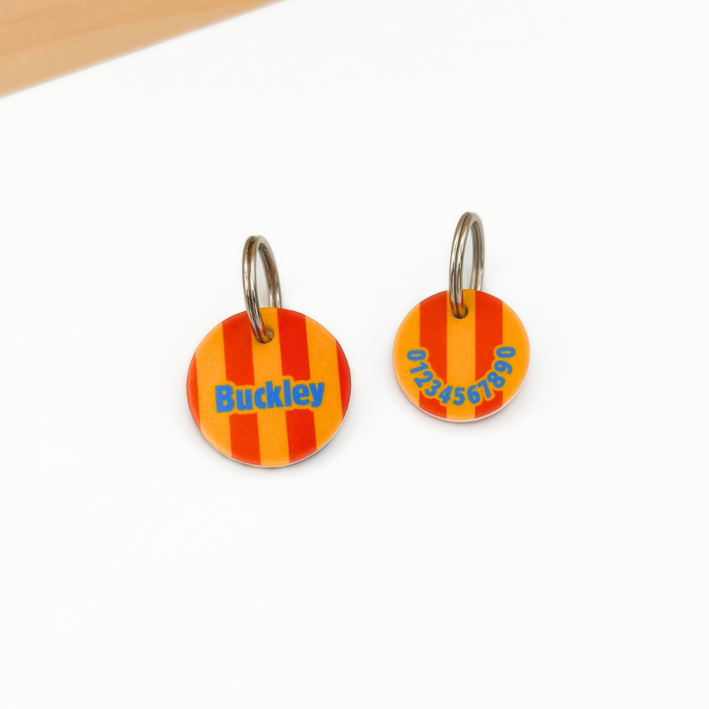 Zwei personalisierte Acryl-Hundemarken mit orange-gelbem Streifenmuster liegen auf einem weißen Untergrund. Die linke Marke zeigt den Namen „Buckley“, die rechte die Rückseite mit Telefonnummer. Beide Hundemarken sind mit silberfarbenen Metallringen versehen. Oben im Bild ist eine helle Holzleiste sichtbar.