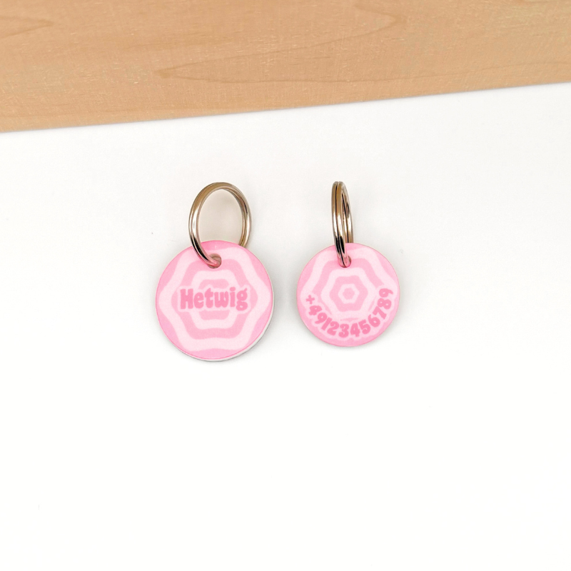 Eine große und eine kleine Hundemarke mit einem verspielten mädchenhaften Design in pink mit Blumen Muster liegen auf einem weißen Boden. Sie haben Kontaktdaten graviert bzw. bedruckt