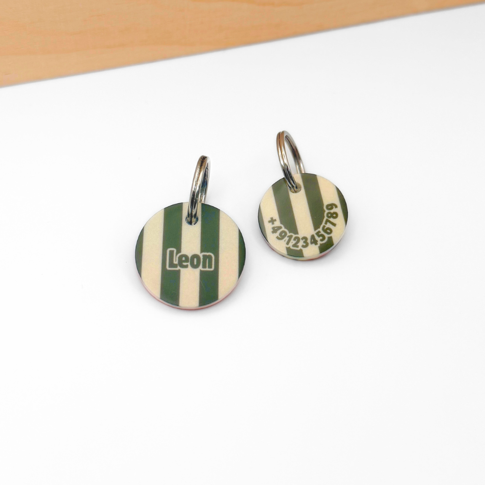Zwei personalisierte Hundemarken aus Acryl mit khaki-beigen Streifen liegen auf weißem Untergrund. Links befindet sich die Vorderseite mit dem Namen „Leon“, rechts die Rückseite mit Telefonnummer. Die Marken sind an silberfarbenen Metallringen befestigt. Im oberen Bildbereich verläuft eine helle Holzleiste.