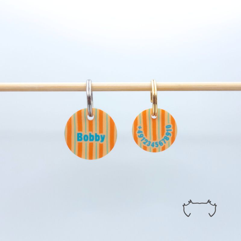 Vorder- und Rückseite einer personalisierten Acryl-Hundemarke für Hunde an einer hellen Stange, vorne mit Name „Bobby“, hinten mit Telefonnummer, beide Seiten im orange-türkisen Streifenmuster.