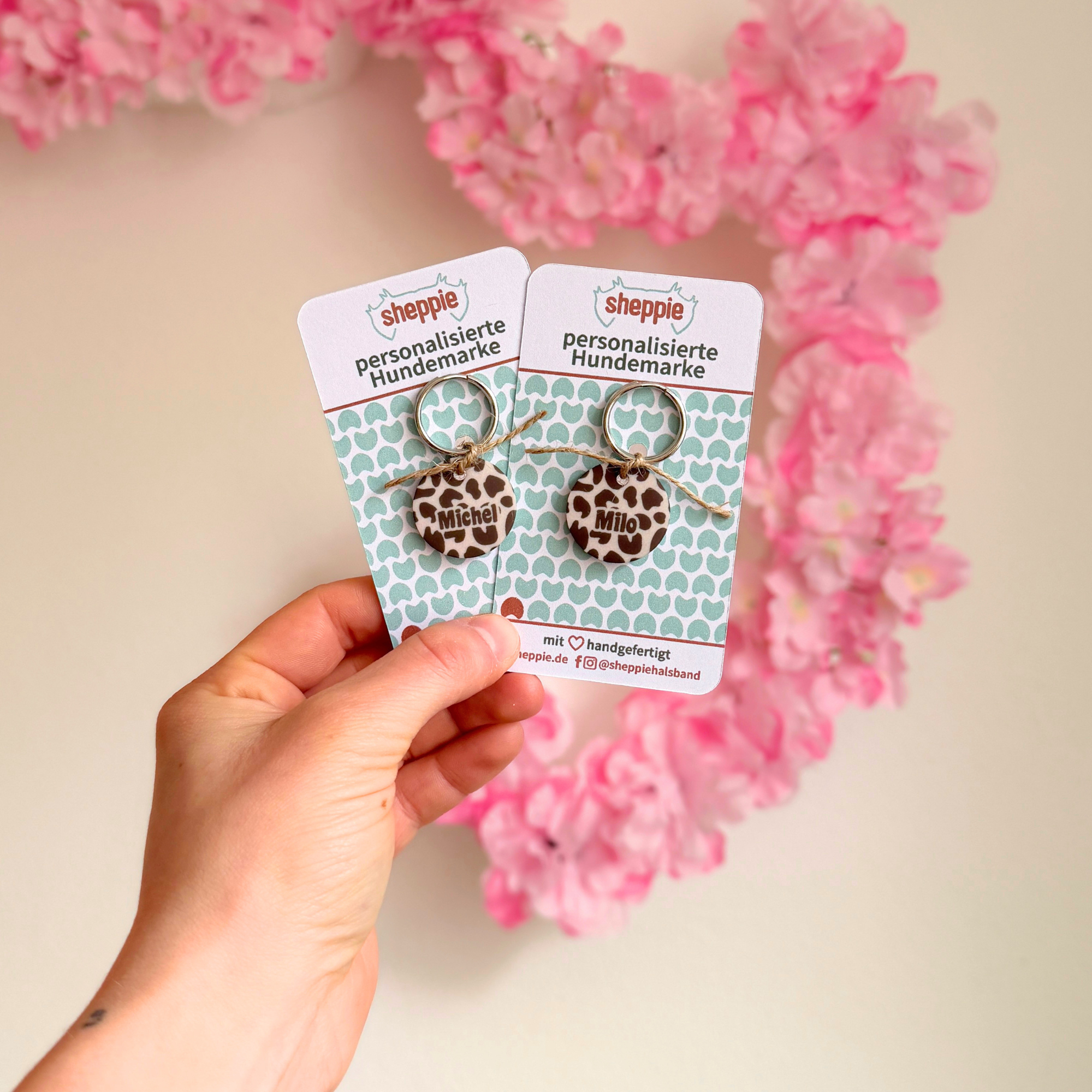 Hand präsentiert personalisierte Acryl Hundemarken im Leo Design auf Produktkarten vor rosa Hintergrund