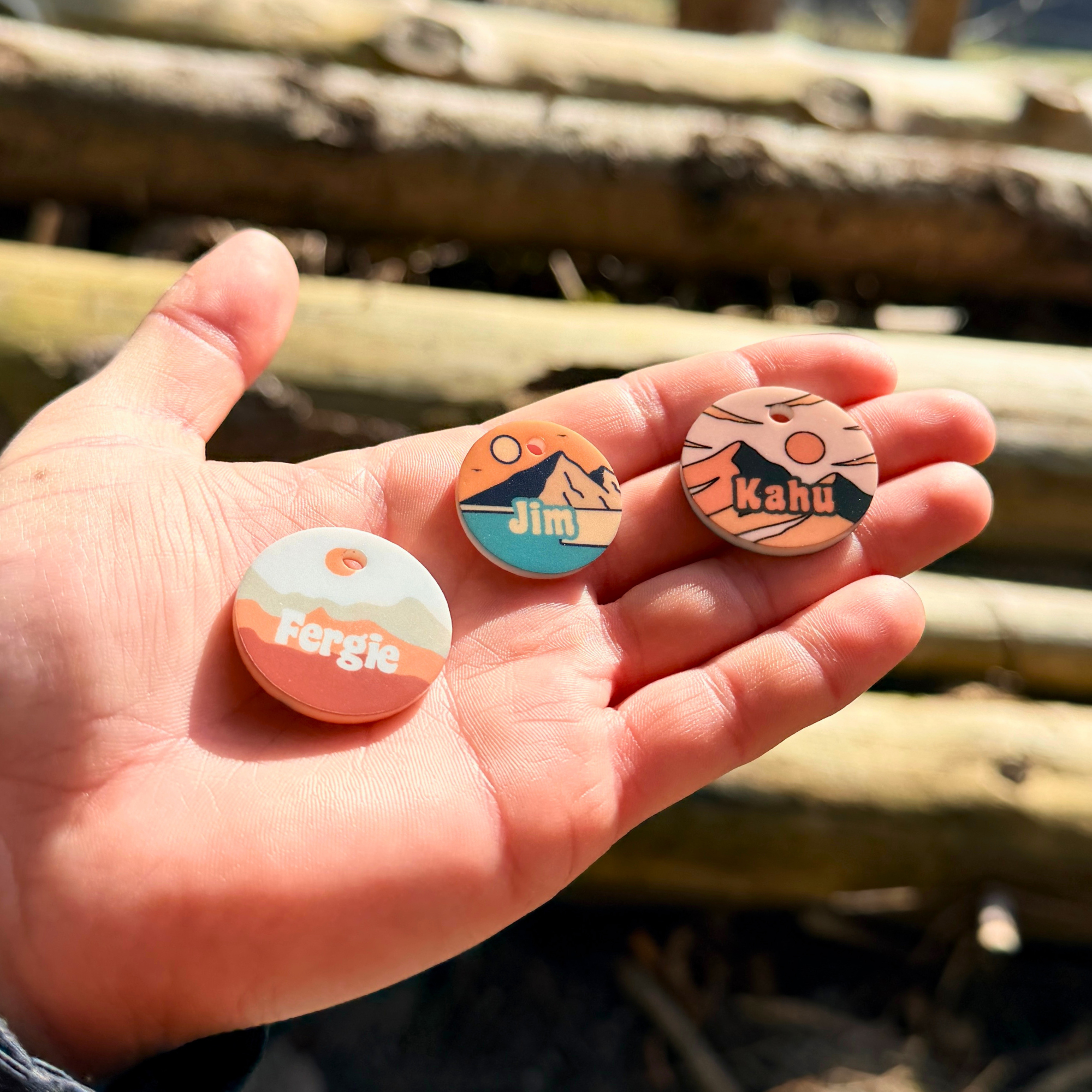 Hand hält zwei personalisierte Acryl Hundemarken mit Berg- und Naturmotiven vor Holzstapel im Hintergrund